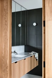 Modern minimalist bathroom interior styled by Bek Sheppard with wooden vanity, dark tiled wall, and white basin viewed through doorway.