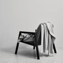 Black armchair with a draped grey knit blanket against a plain grey wall and speckled floor