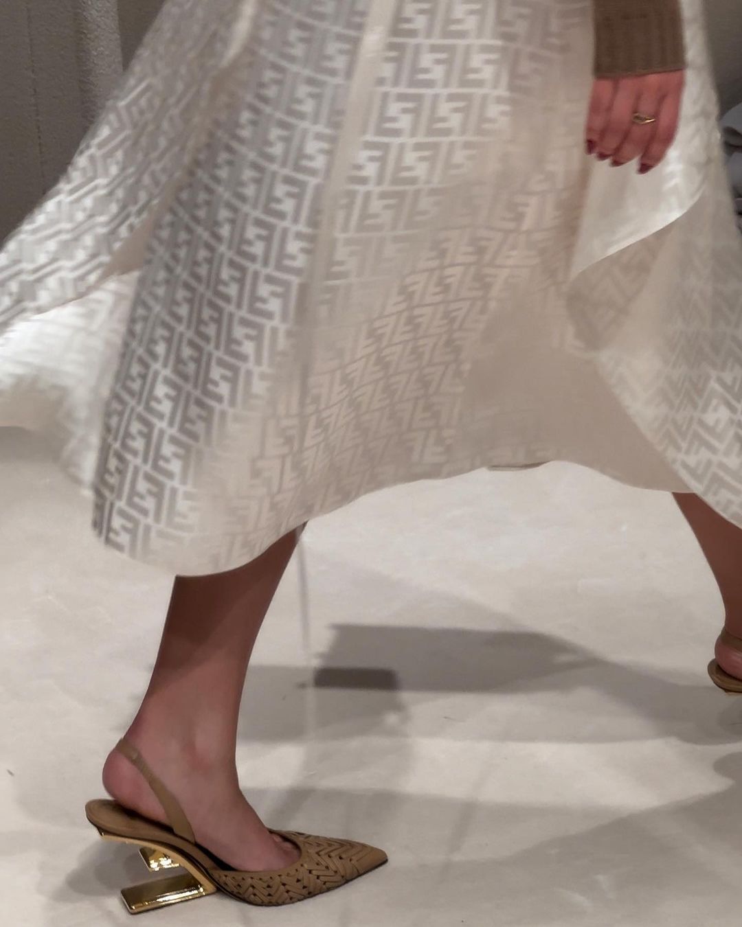 Runway shot showcases a tan pointed-toe heel with a gold architectural heel under a flowing white Fendi logo-patterned dress.