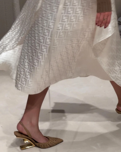 Runway shot showcases a tan pointed-toe heel with a gold architectural heel under a flowing white Fendi logo-patterned dress.