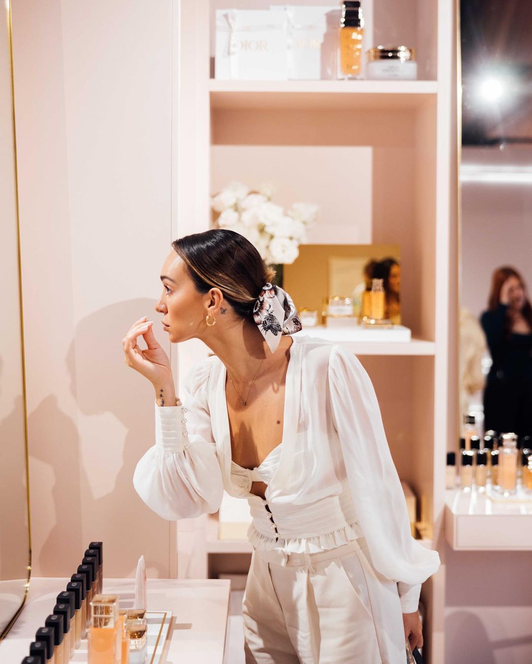 Woman applying makeup in a beauty store, wearing a white top, with beauty products displayed on shelves and a makeup counter.