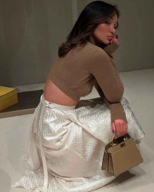 Young woman poses in a brown sweater and patterned white skirt, holding a beige handbag in an indoor setting with neutral tones.