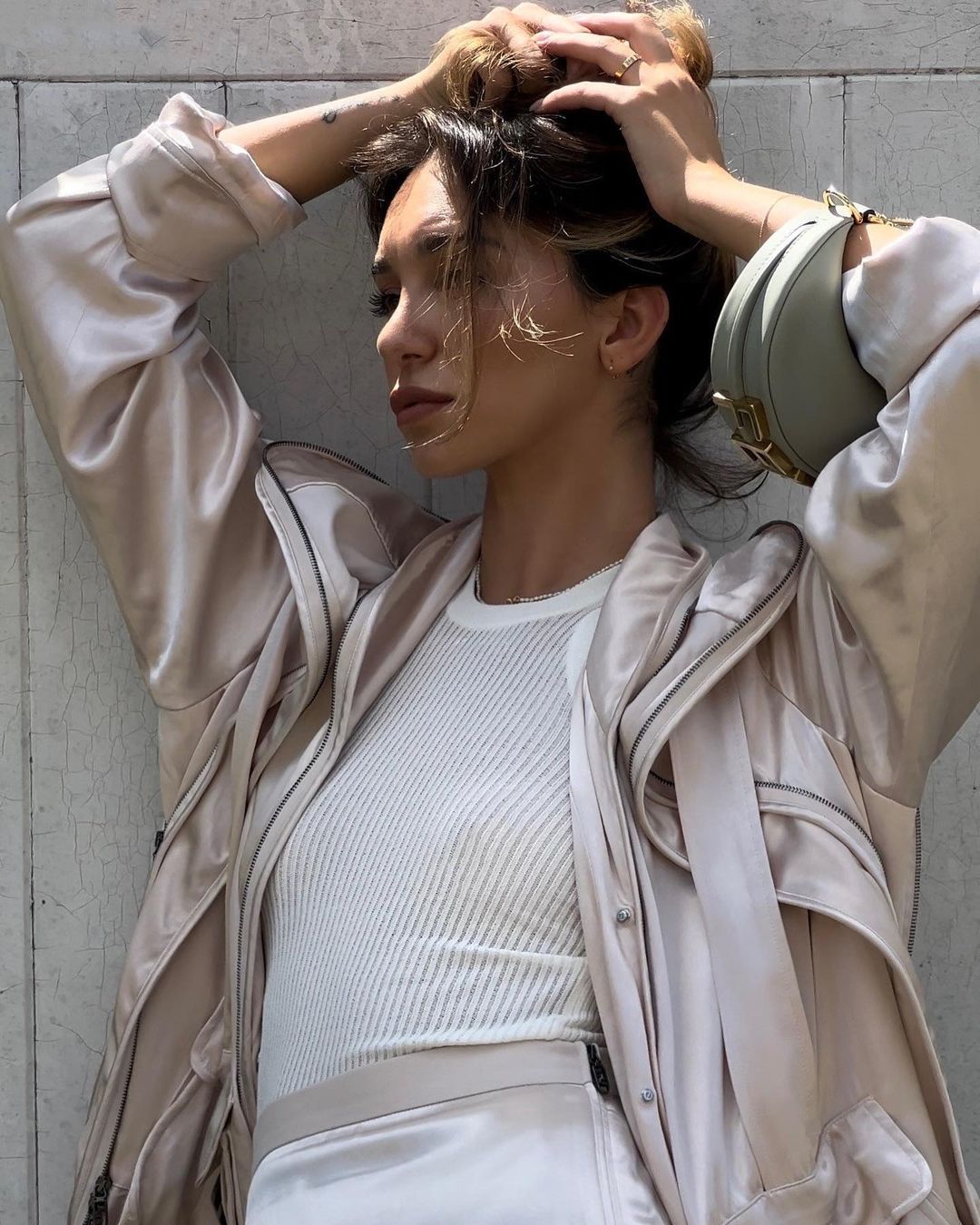 Stylish woman in neutral-toned matching set, silk coat with bag slung over shoulder against pale concrete wall, putting her hair up.