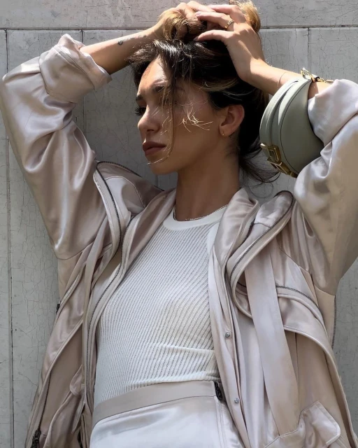 Stylish woman in neutral-toned matching set, silk coat with bag slung over shoulder against pale concrete wall, putting her hair up.