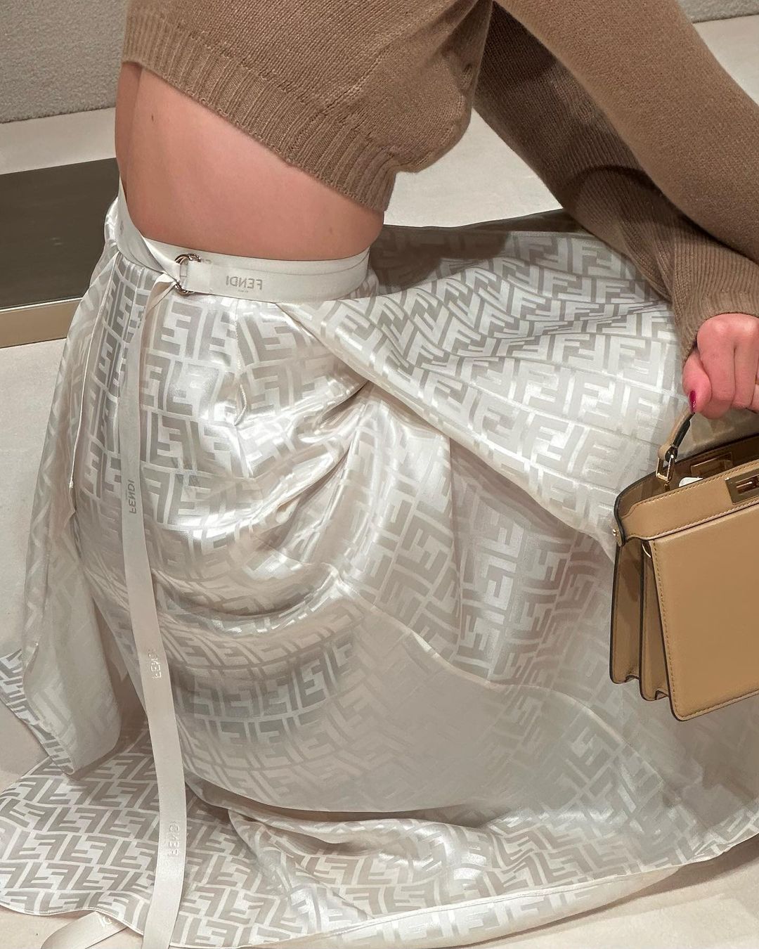 Woman wearing a brown sweater and Fendi patterned skirt with matching handbag, seated indoors with neutral toned background.
