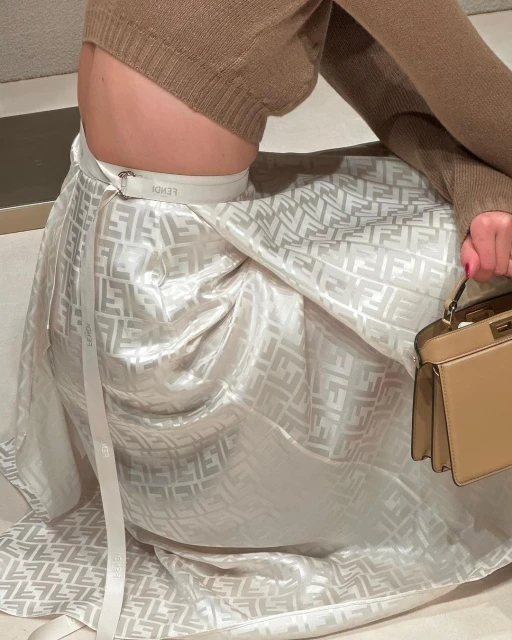 Woman wearing a brown sweater and Fendi patterned skirt with matching handbag, seated indoors with neutral toned background.