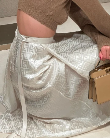 Woman wearing a brown sweater and Fendi patterned skirt with matching handbag, seated indoors with neutral toned background.