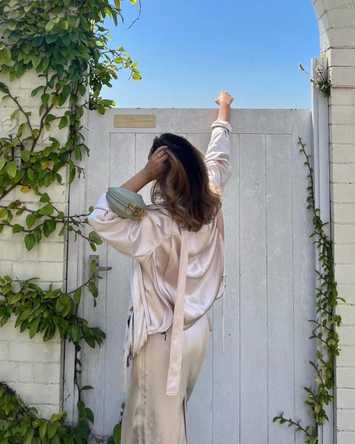 Adult woman stands outdoors in a champagne colored skirt and top holding onto a white gate surrounded by green vines against a blue sky.