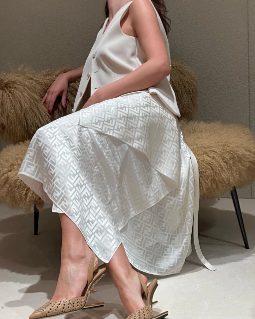 Woman in a cream vest and Fendi-patterned skirt sitting on a fuzzy chair, wearing decorative heels. Neutral color palette indoors.