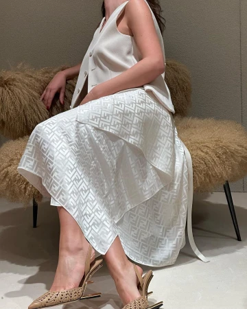 Woman in a cream vest and Fendi-patterned skirt sitting on a fuzzy chair, wearing decorative heels. Neutral color palette indoors.