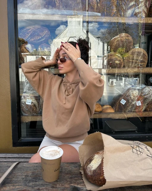Woman in a sweatshirt sitting at a table with coffee and bread, a bakery is visible behind her.