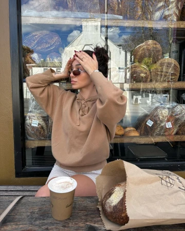 Woman in a sweatshirt sitting at a table with coffee and bread, a bakery is visible behind her.