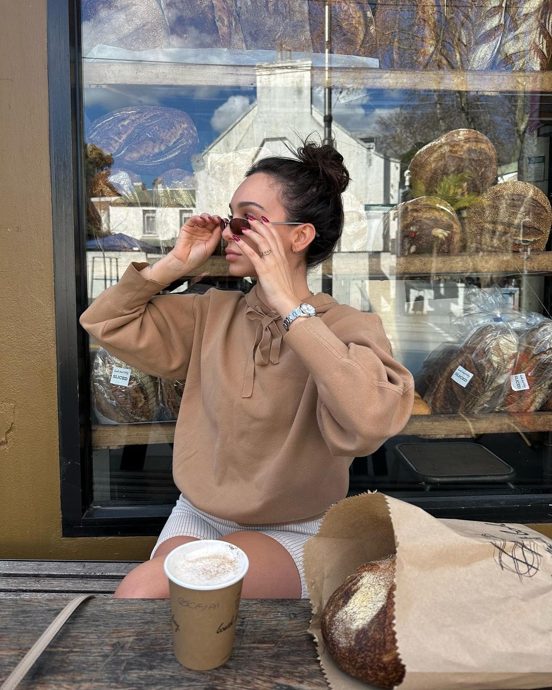Woman in casual attire adjusts sunglasses outside a bakery with bread displayed in the window and a coffee cup on the table.