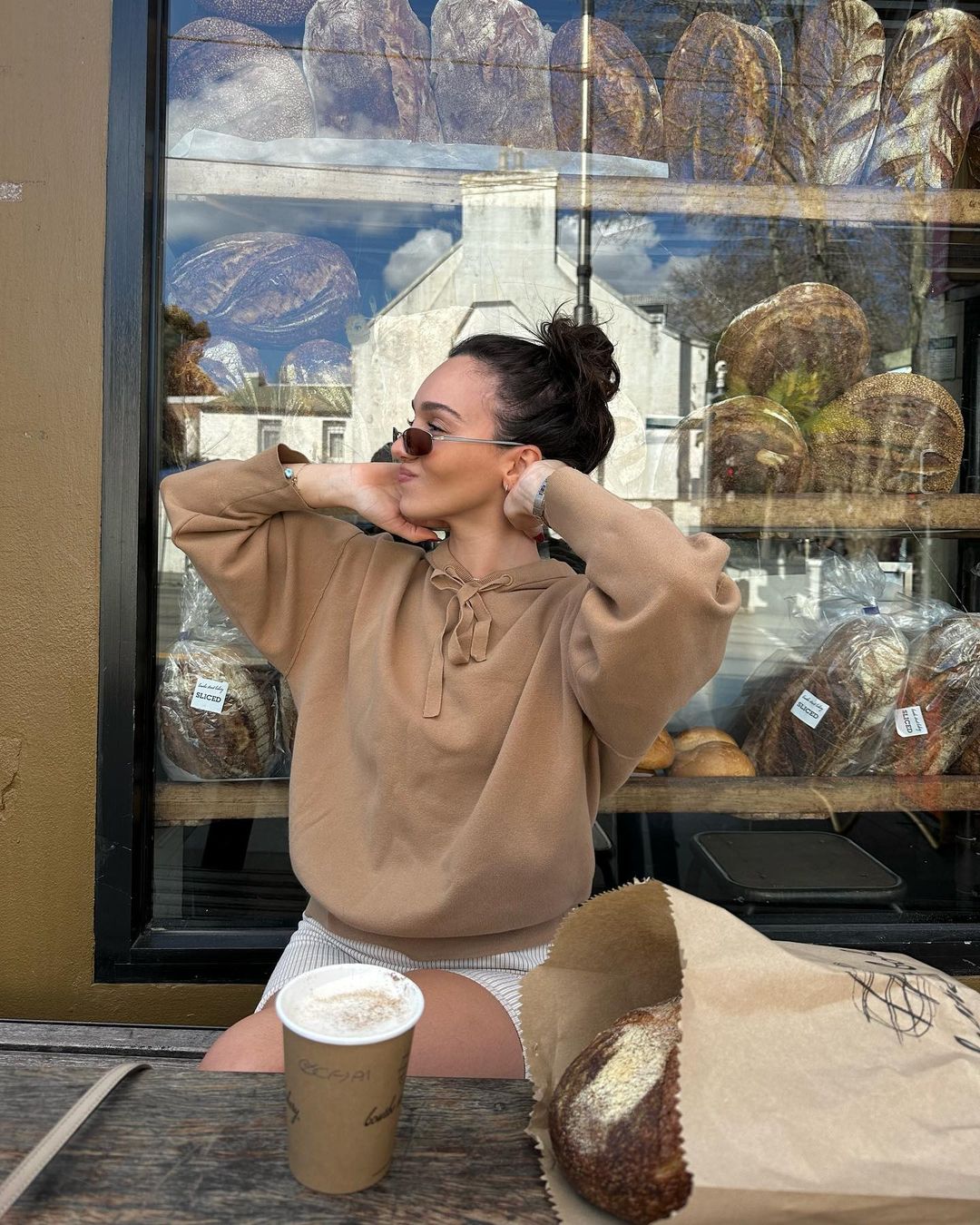 Woman in tan hoodie poses near bakery window with bread, coffee, and paper bag on wooden table, in daylight, outdoor urban setting.