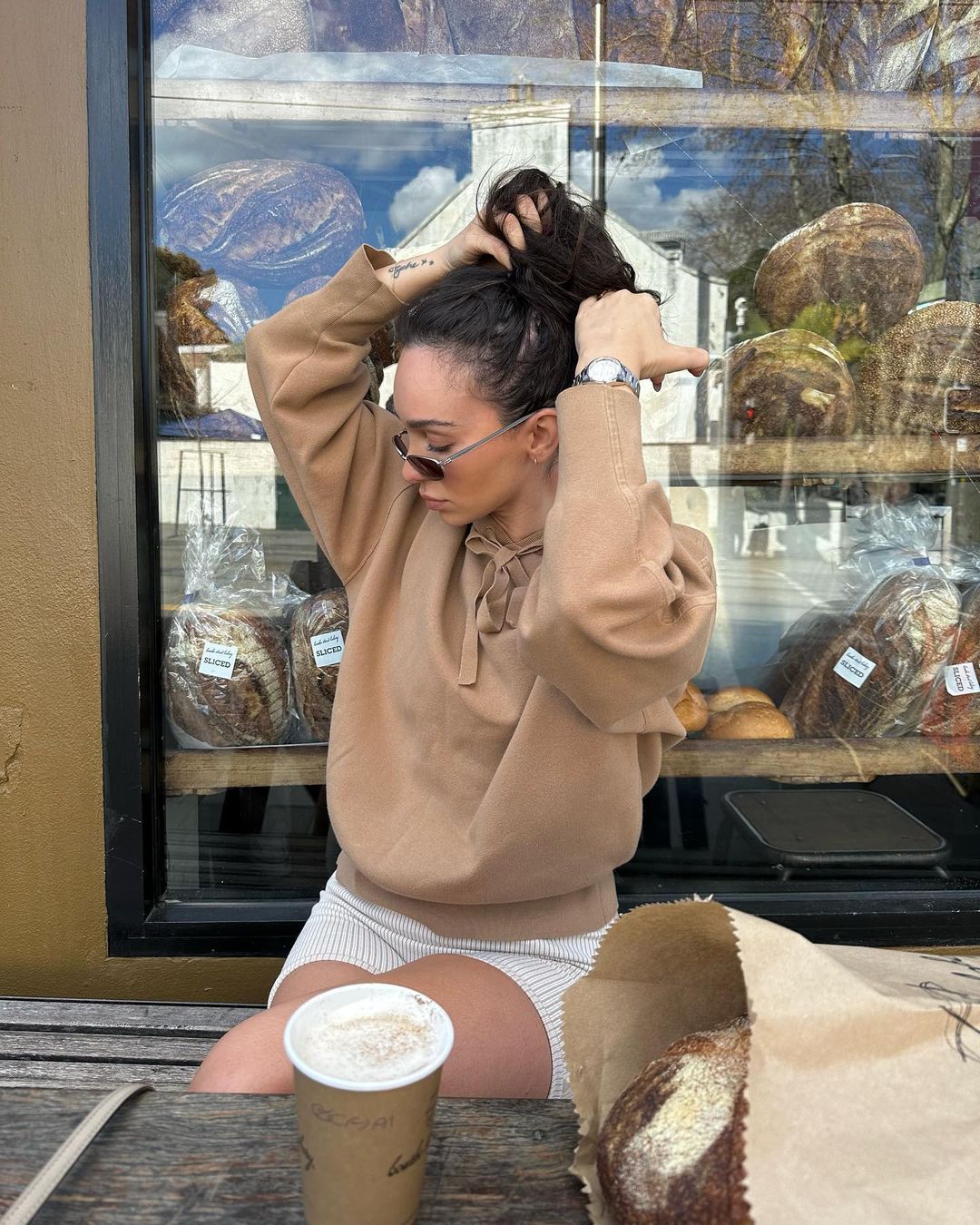 Brunette woman in a tan hoodie and shorts sits on a bench outside, tying her hair up, with coffee and bread in view.
