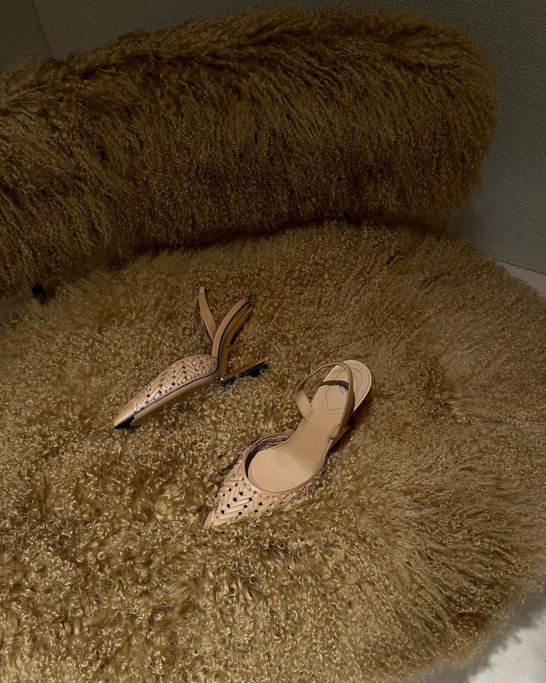 Two light-brown heeled shoes sit on a furry, round rug. The shoes are pointed-toe with intricate cut-out patterns and slingback straps.