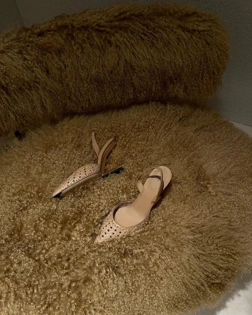 Two light-brown heeled shoes sit on a furry, round rug. The shoes are pointed-toe with intricate cut-out patterns and slingback straps.
