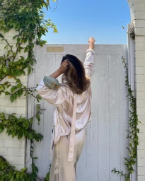 Woman in satin outfit with shoulder bag reaching up toward a white wooden gate framed by brick and ivy.