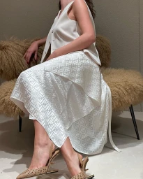 Woman seated on a furry bench wearing a white vest and patterned white skirt with beige slingback heels.