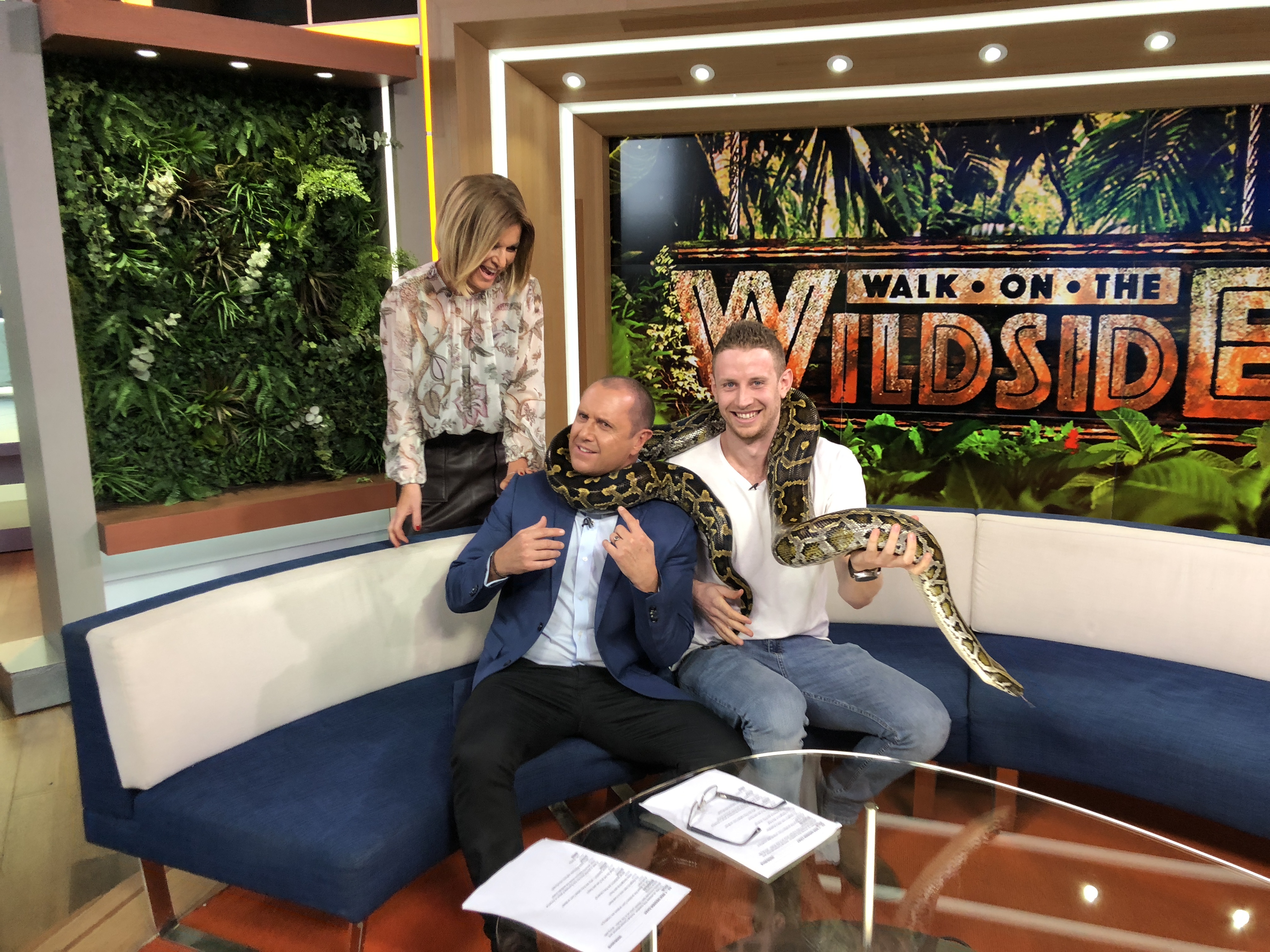 Three people pose on a couch with a snake draped around two of their necks, in a bright studio setting with jungle-themed backdrop.