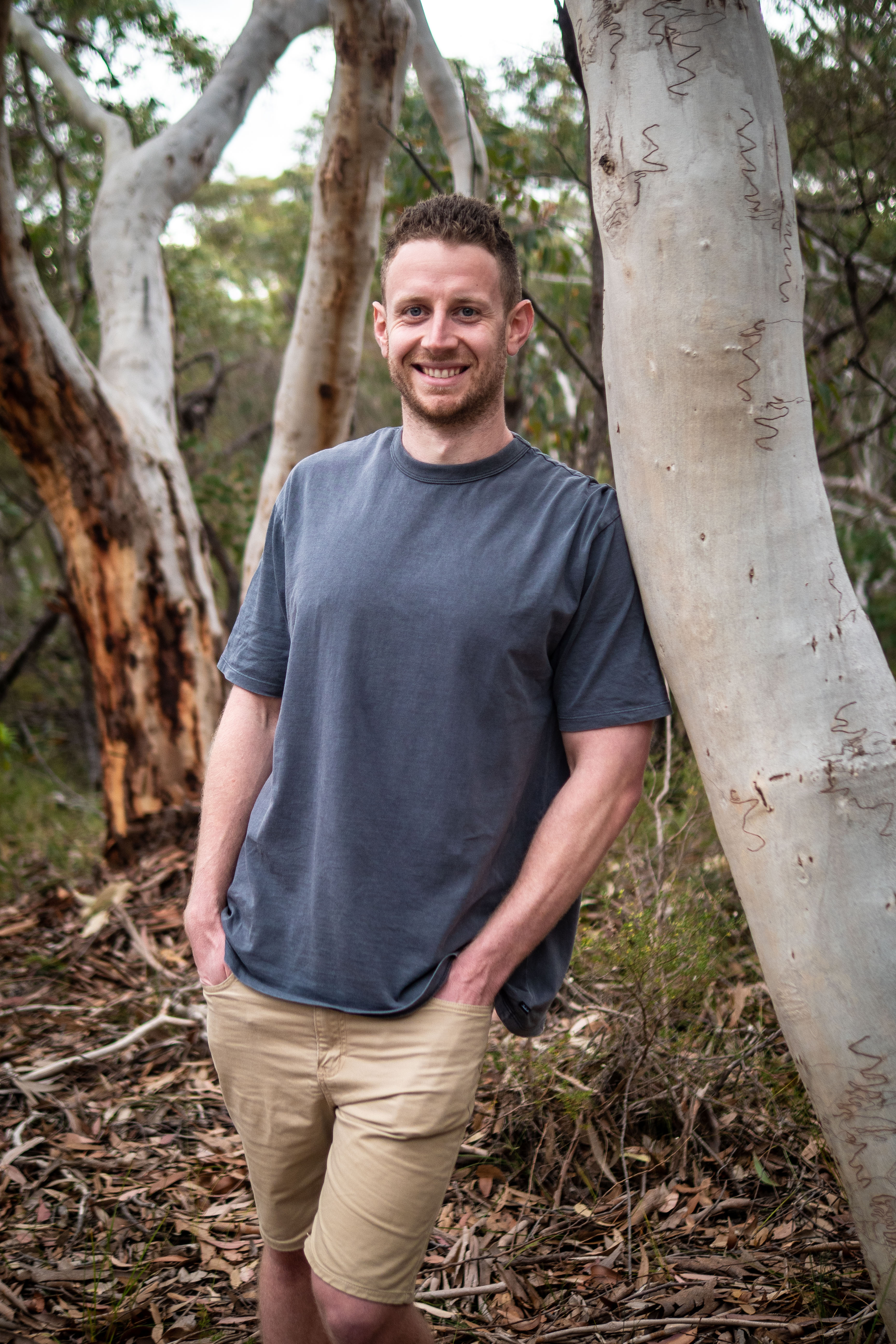 Man in grey t-shirt and khaki shorts stands against a eucalyptus tree smiling in an outdoor woodland setting with dappled lighting.