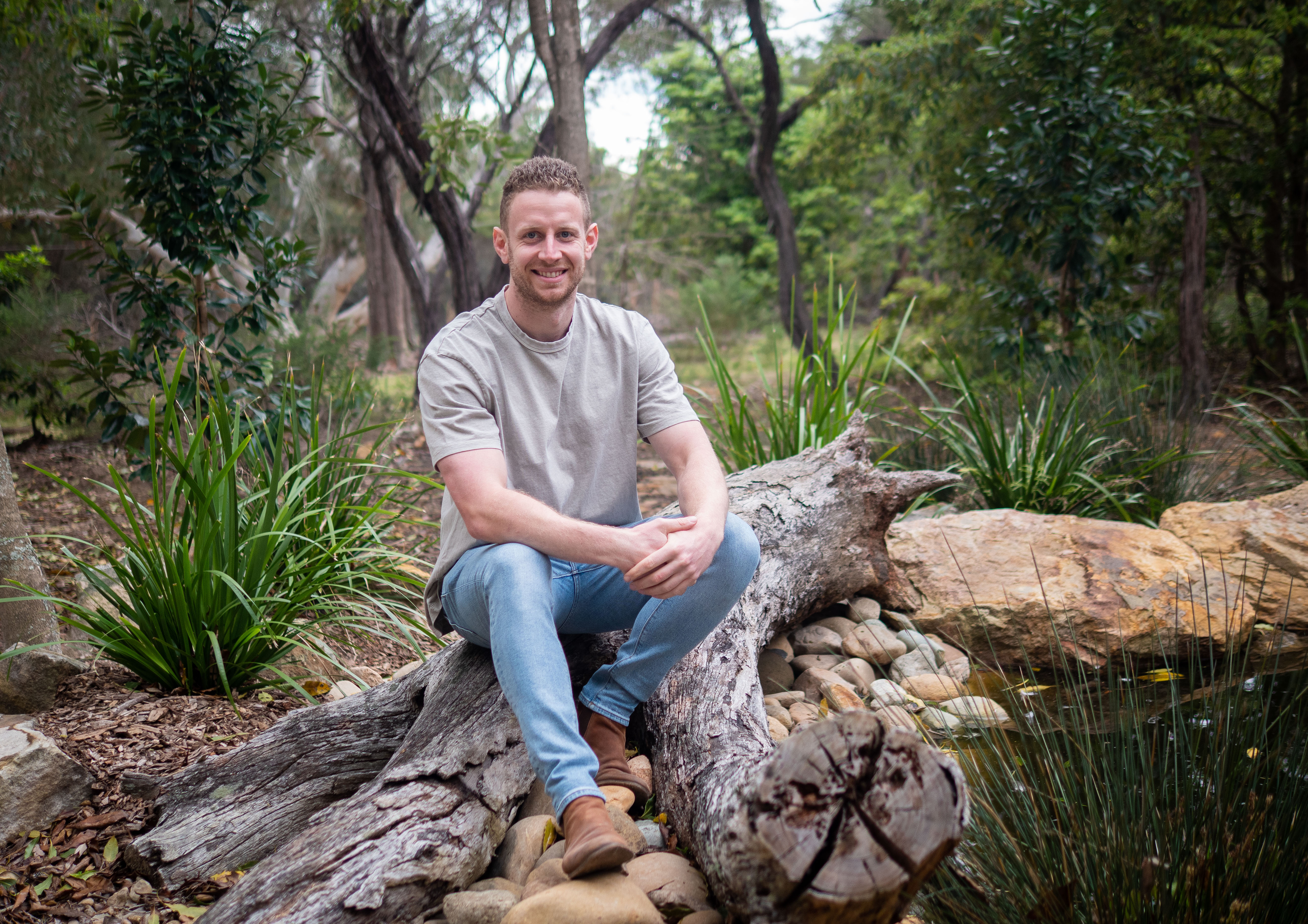 A smiling adult male sits on a fallen log in a forest with lush green foliage, wearing a t-shirt, light blue jeans, and tan boots.
