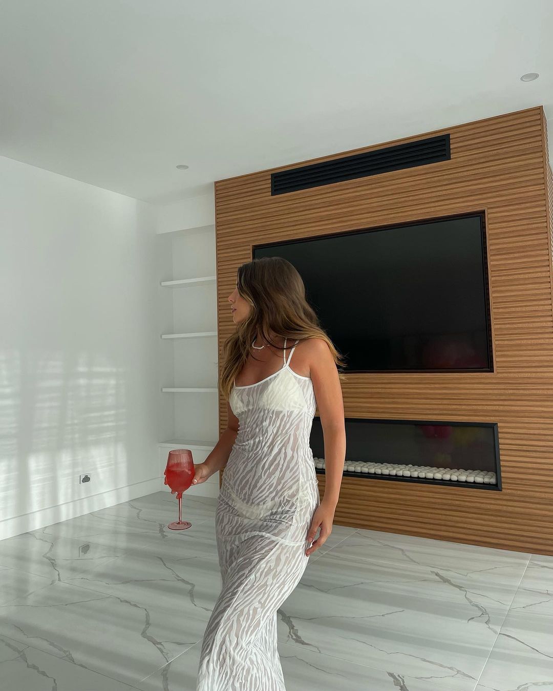 Woman in sheer white dress holds a pink drink in an indoor setting with wood paneling and white walls, looking away from the camera.