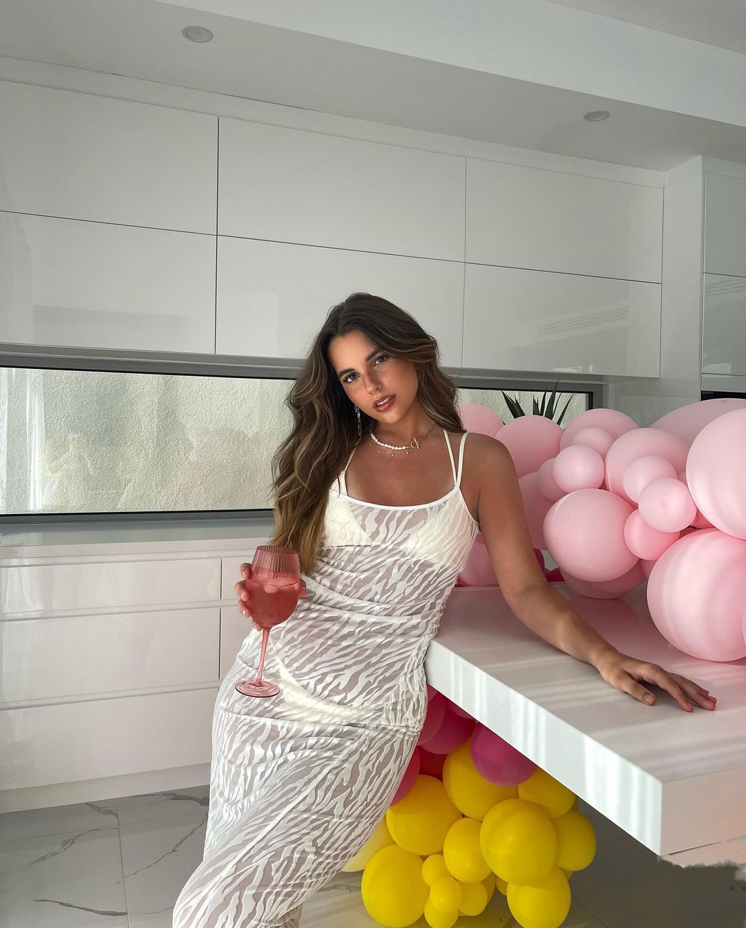 Woman in a zebra-striped dress stands next to a kitchen counter, holding a pink drink, surrounded by pink and yellow balloons.
