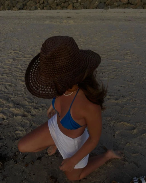 Woman in a blue bikini and white shorts kneeling on the sand at the beach wearing a brown woven sun hat in an eye-level shot.