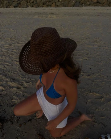 Woman in a blue bikini and white shorts kneeling on the sand at the beach wearing a brown woven sun hat in an eye-level shot.