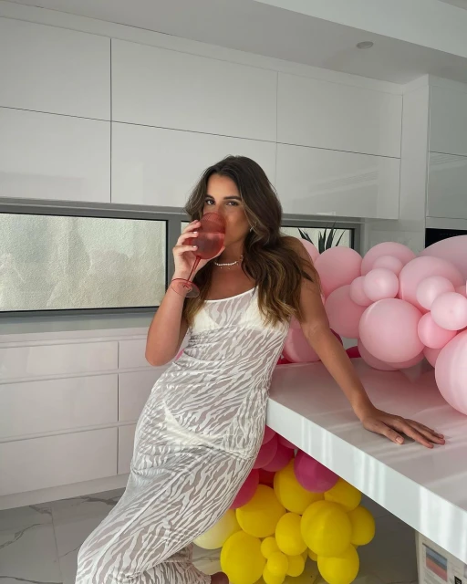 Woman in a zebra print dress leans on a white countertop, drinking from a stemmed glass. Balloons and white cabinetry visible.
