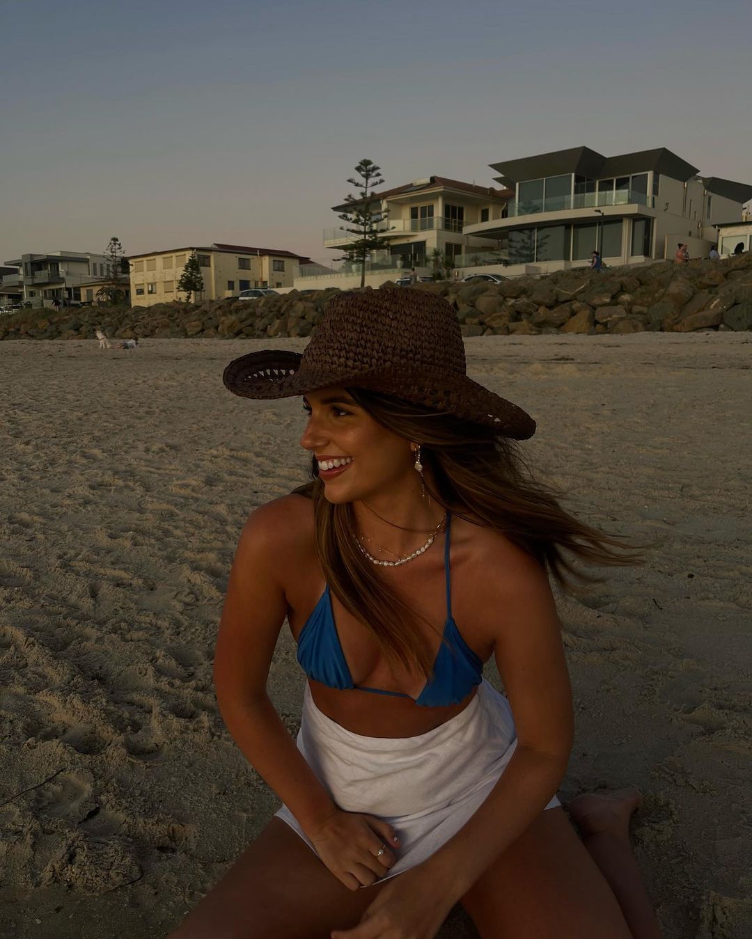 Young woman in a blue bikini top and white skirt wearing a brown hat on the sandy beach, smiling with houses in the background.