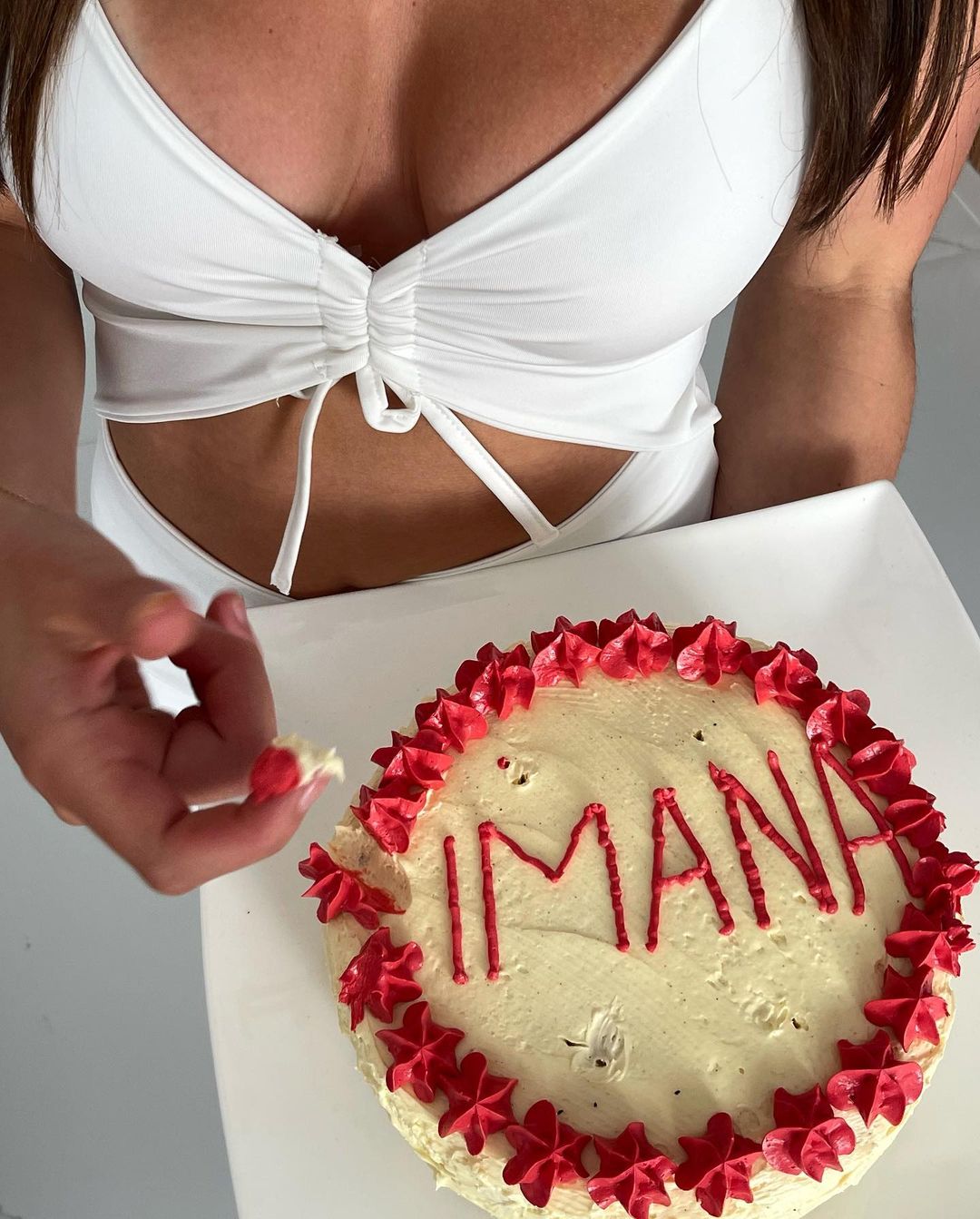Woman holding a cake with 'IMANA' written in red icing, featuring white frosting and red icing stars on a square white plate.