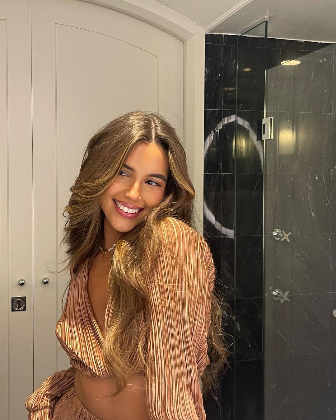 Smiling brunette woman with long wavy hair wearing a peach-colored blouse with marble tiled shower and white door in background.