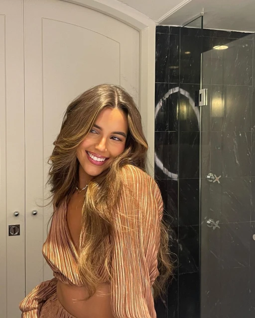 Smiling brunette woman with long wavy hair wearing a peach-colored blouse with marble tiled shower and white door in background.