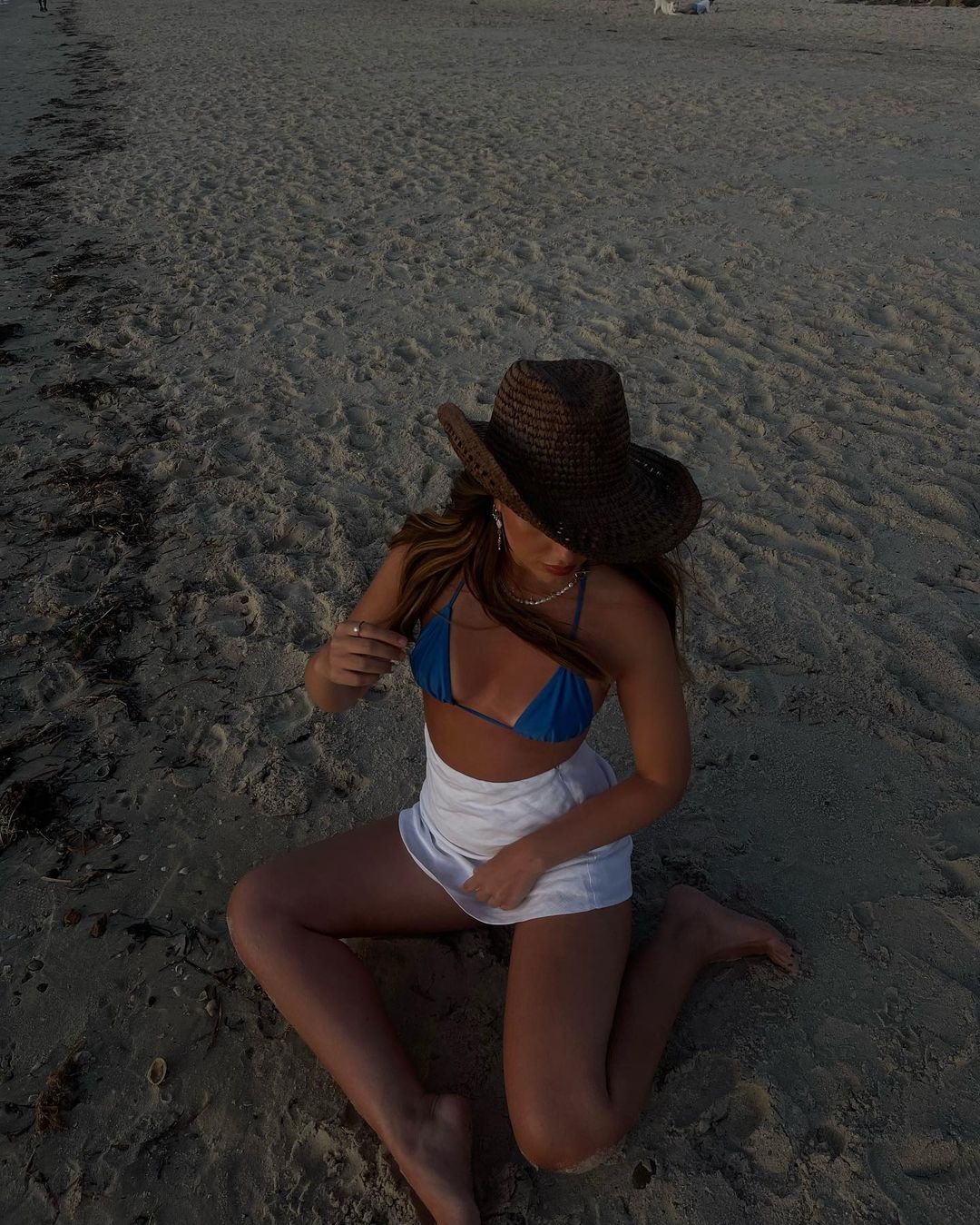 Young woman in a bikini and skirt poses on a sandy beach wearing a cowboy hat, enjoying the sunshine in a relaxed outdoor setting.