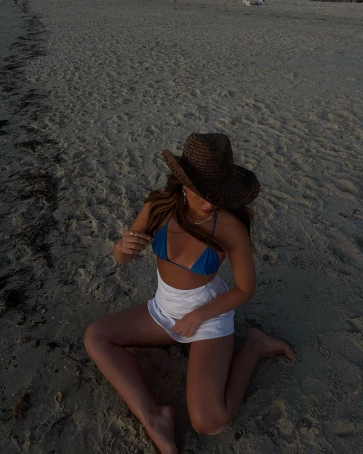 Young woman in a bikini and skirt poses on a sandy beach wearing a cowboy hat, enjoying the sunshine in a relaxed outdoor setting.