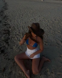 Woman wearing a blue bikini, white mini skirt, and straw hat kneeling on a sandy beach.