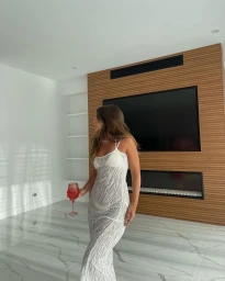 Woman in a sheer patterned maxi dress holding a red glass inside a modern living room with wood paneling.