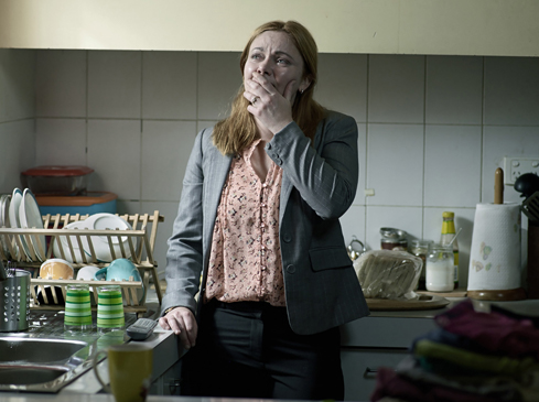 Adult woman stands in a kitchen with a concerned expression, wearing a blazer over a floral top. A dish rack and sink are visible.