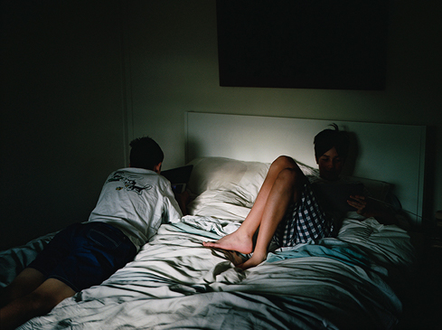 Two people, one reclining and one seated, are captured indoors on a bed, bathed in a mix of natural and artificial light, creating a moody atmosphere.