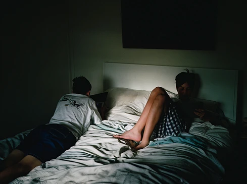 Two people, one reclining and one seated, are captured indoors on a bed, bathed in a mix of natural and artificial light, creating a moody atmosphere.