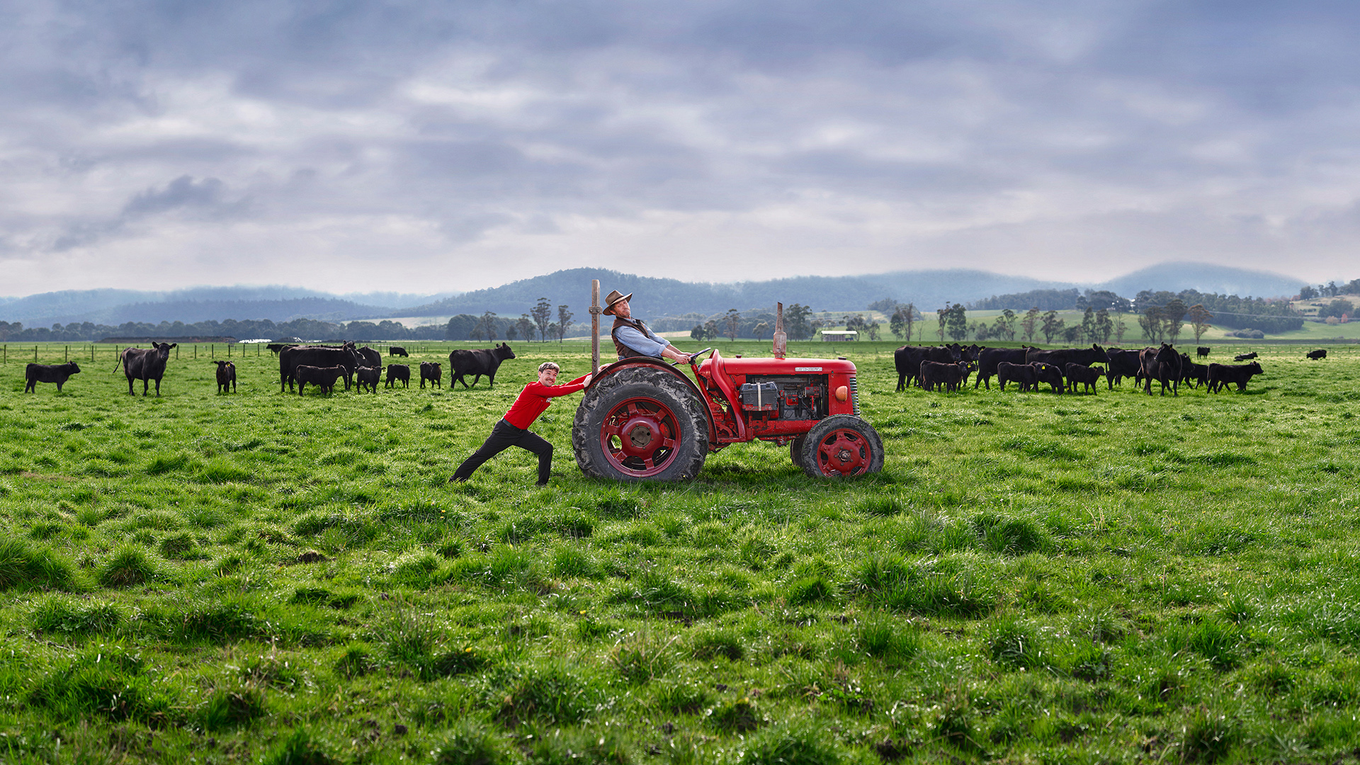 Two people on a rural farm with cattle and green pasture, one pushing a red tractor while the other drives on a cloudy day.