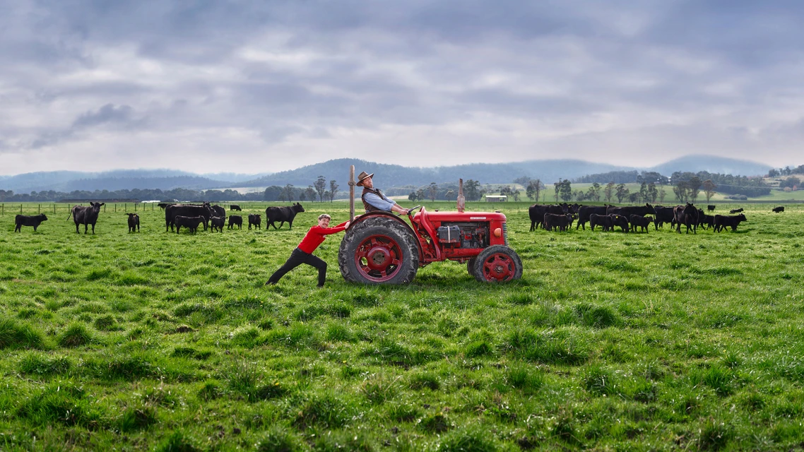 Two people on a rural farm with cattle and green pasture, one pushing a red tractor while the other drives on a cloudy day.
