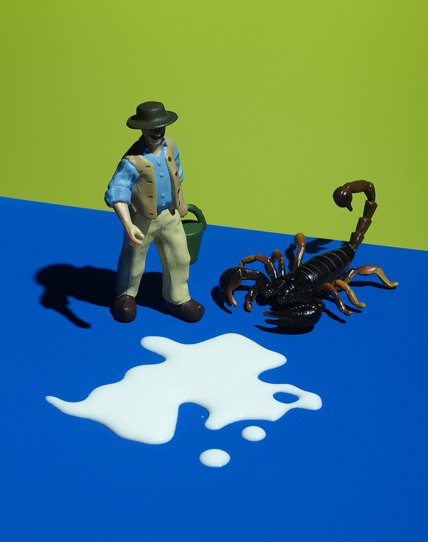 Toy figure of a person with a hat standing next to a toy scorpion with a splash of white liquid on a blue and green surface.
