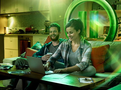 A couple sits on a couch looking at a laptop with a game controller on the coffee table in front of them in a warmly lit living room.