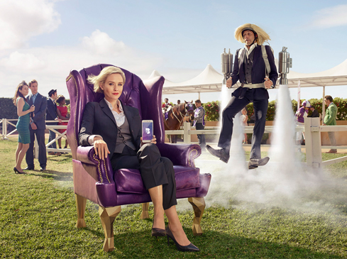 Businesswoman in a purple armchair holds a phone, while a man with a jetpack flies by in an outdoor event, with spectators in the background.