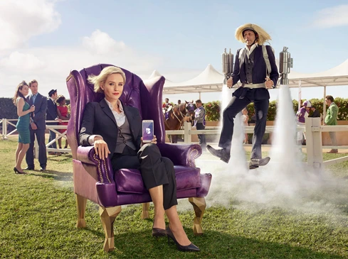 Businesswoman in a purple armchair holds a phone, while a man with a jetpack flies by in an outdoor event, with spectators in the background.