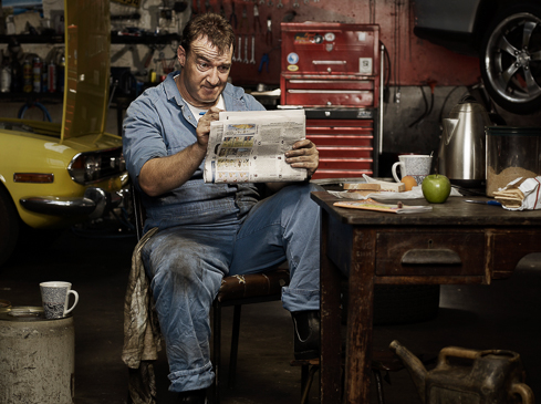 Garage mechanic takes a break, sitting and reading a newspaper next to a yellow car with tools and equipment visible in the shop setting.