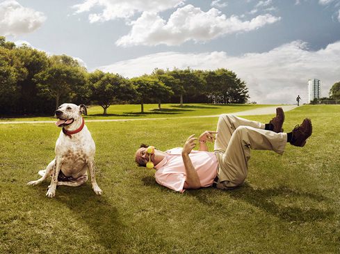Dog sits at attention beside a man lying on his back in a grassy park with tennis balls in his mouth, trees and a building in background.